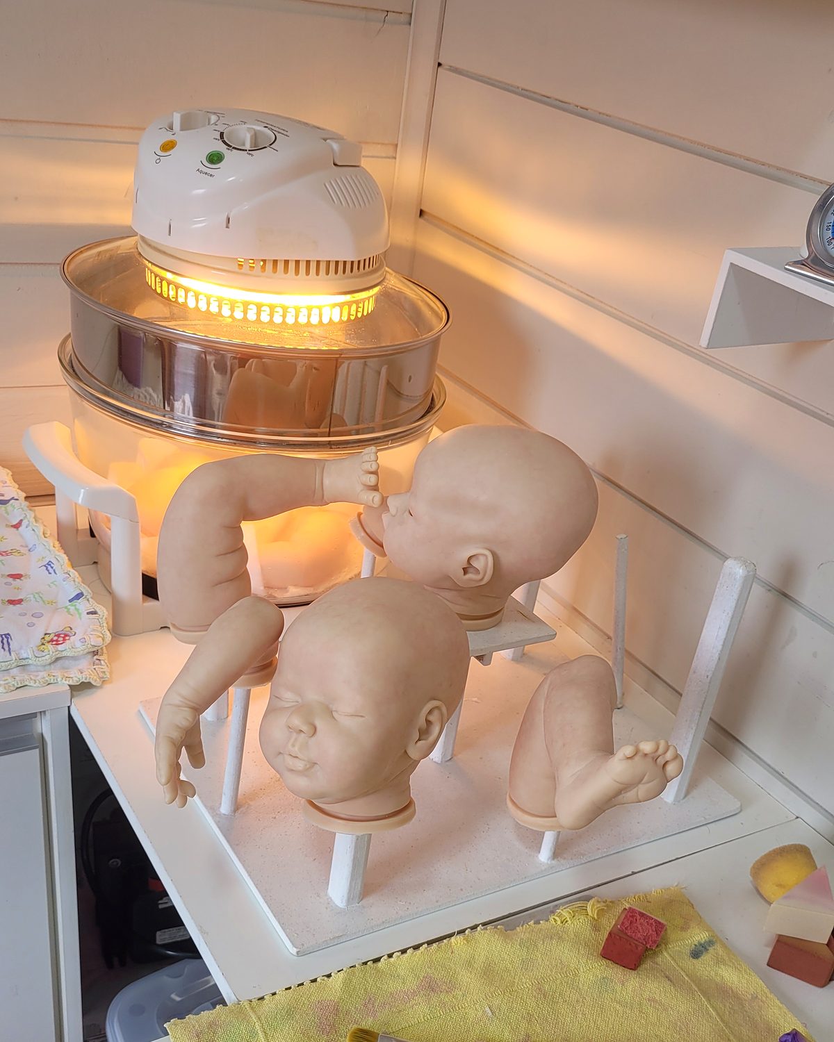 Cabeças e membros de bebê reborn sobre suportes, ao lado do forno do ateliê