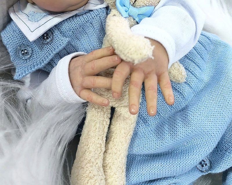 Detalhe das mãos de um bebê reborn segurando um ursinho de pelúcia