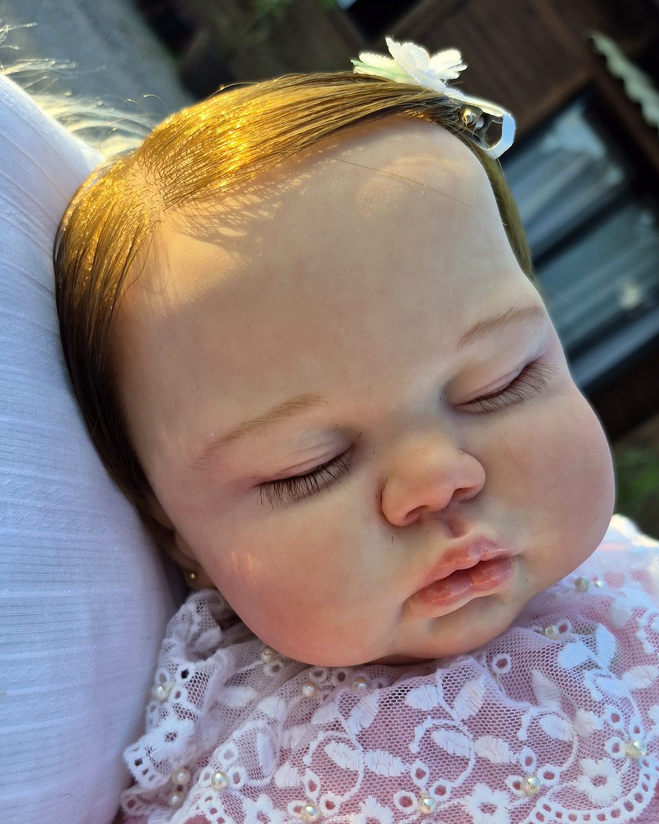 Bebê reborn adormecida, com luz dourada no cabelo, colarinho de renda e expressão serena