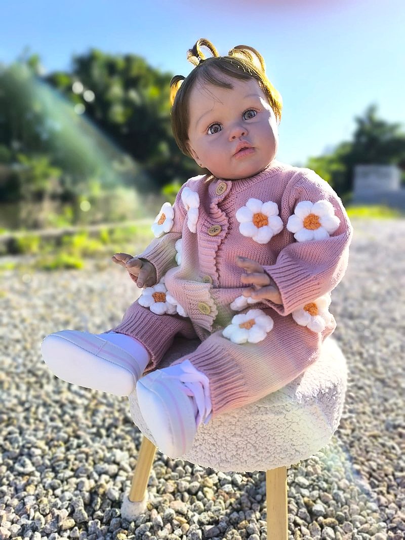Bebê reborn sentada ao ar livre com conjunto de tricô rosa e flores de margarida