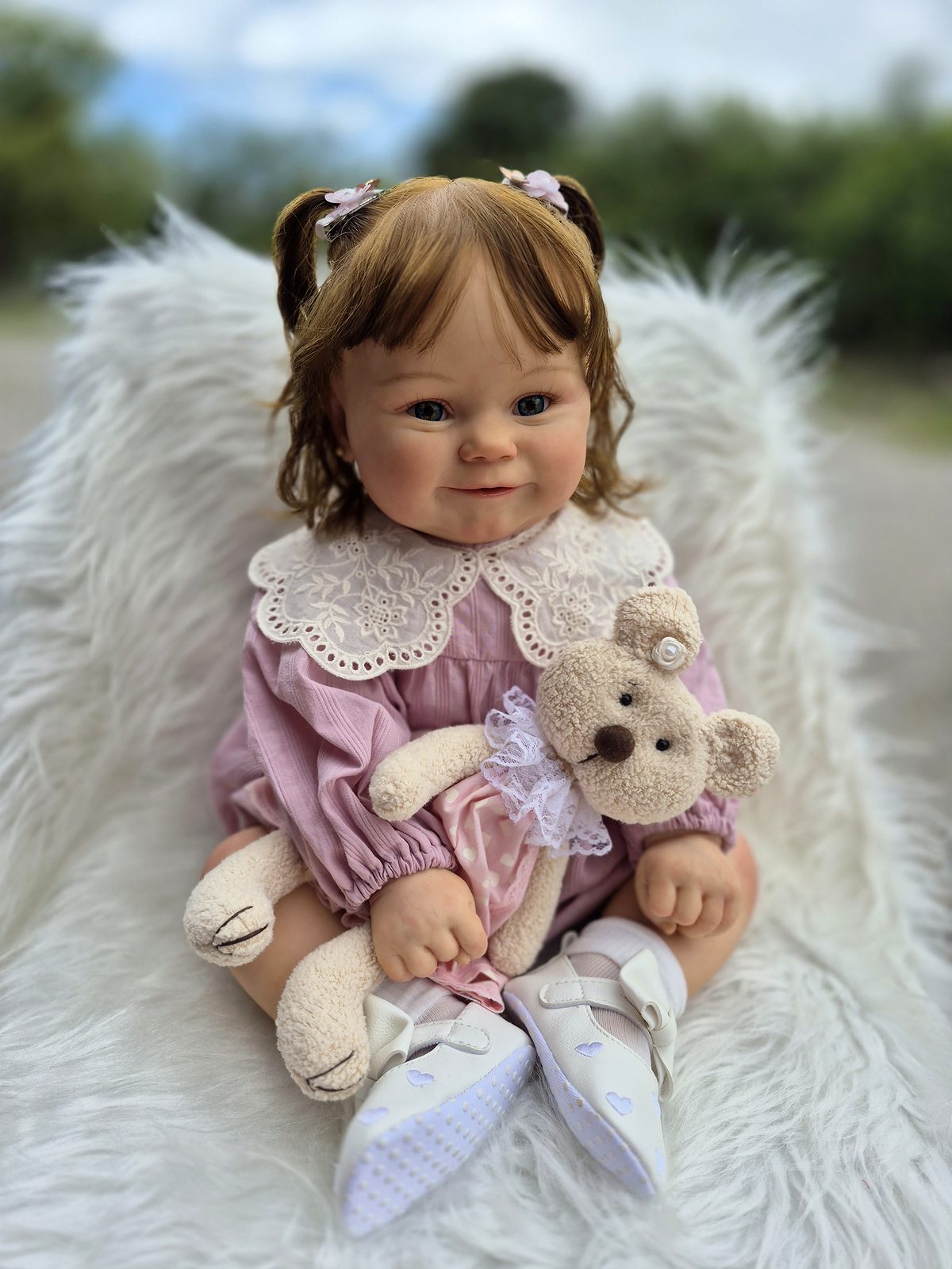 Bebê reborn menina sentada, com maria-chiquinhas, vestido lilás de renda e ursinho de pelúcia