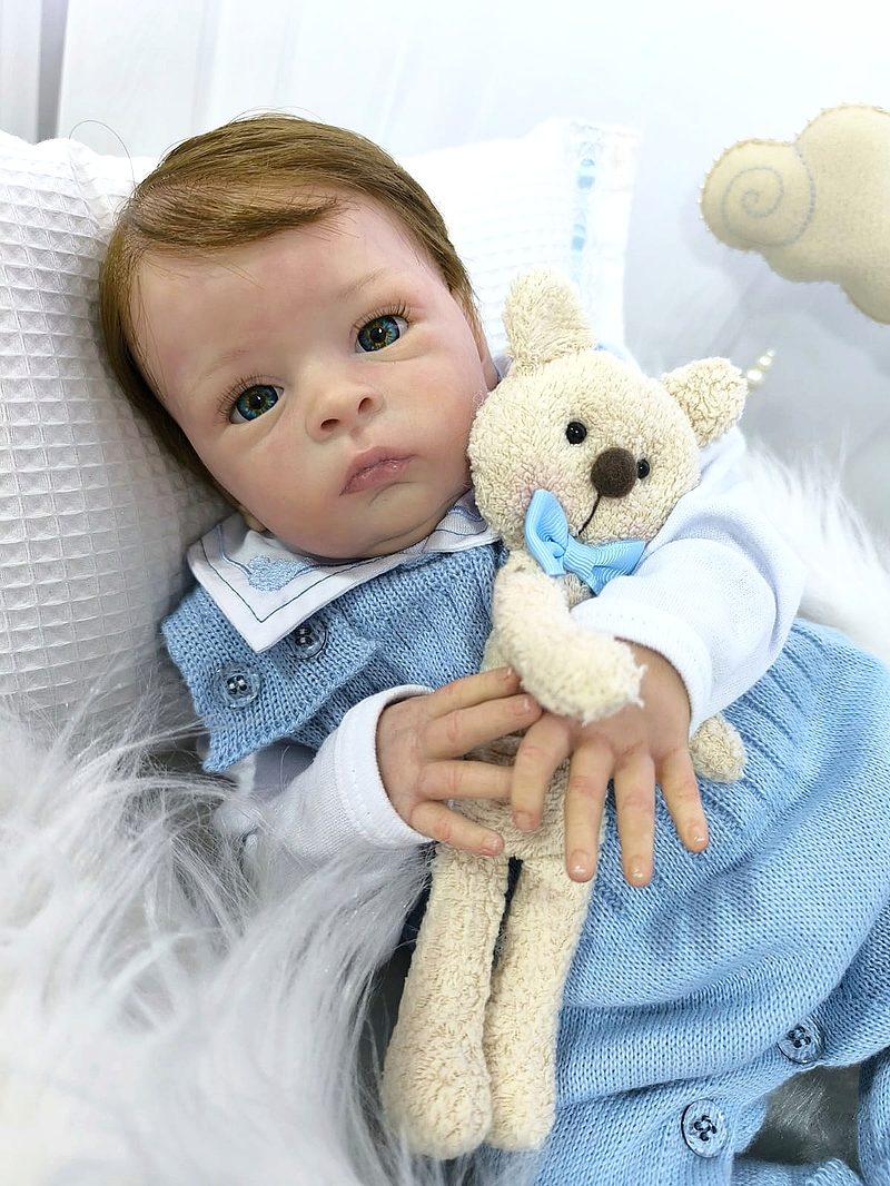 Bebê reborn menino de olhos verdes, conjunto azul de tricô, abraçando um ursinho