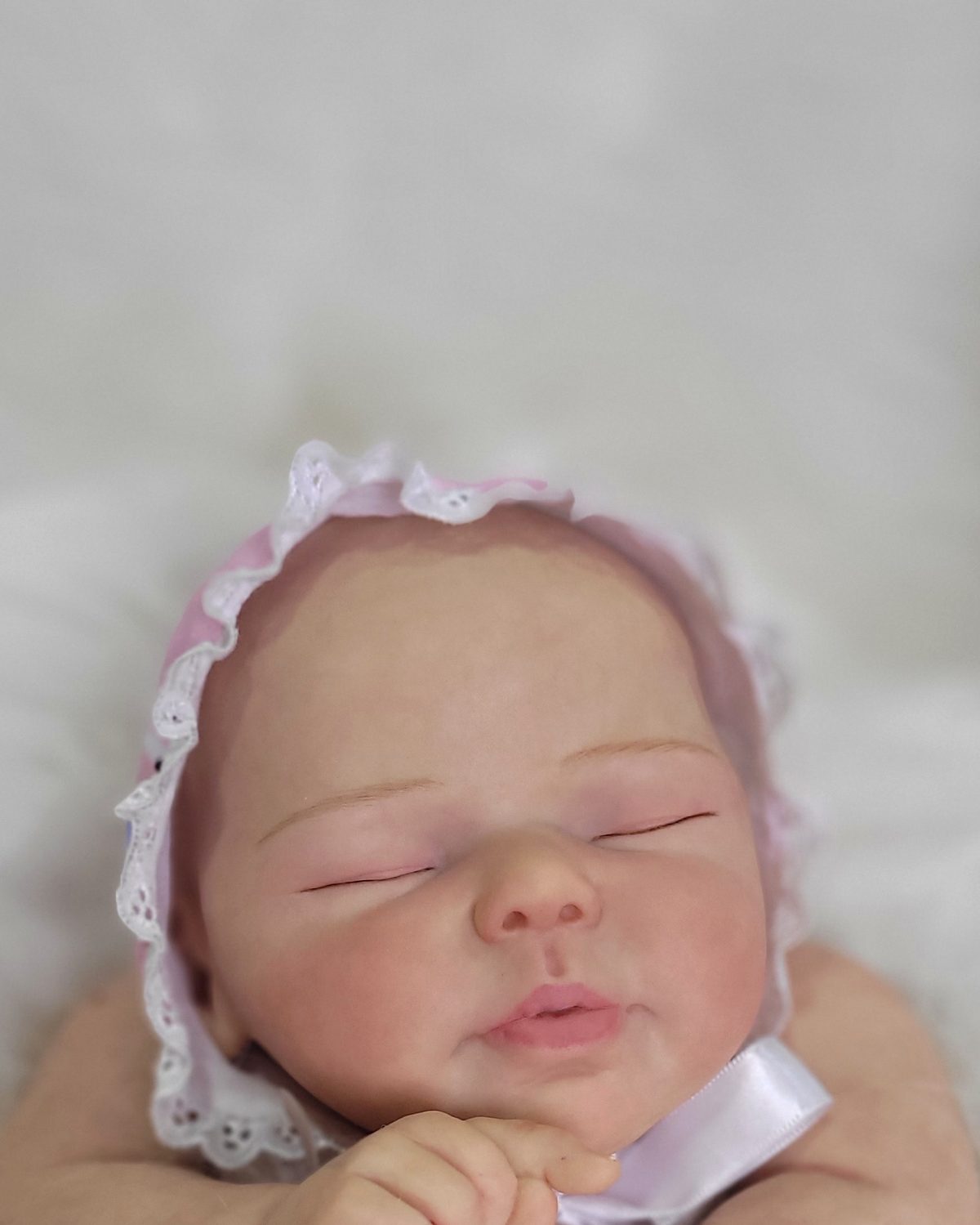Bebê reborn recém-nascida adormecida com touca rosa de renda, mãos juntas junto ao rosto